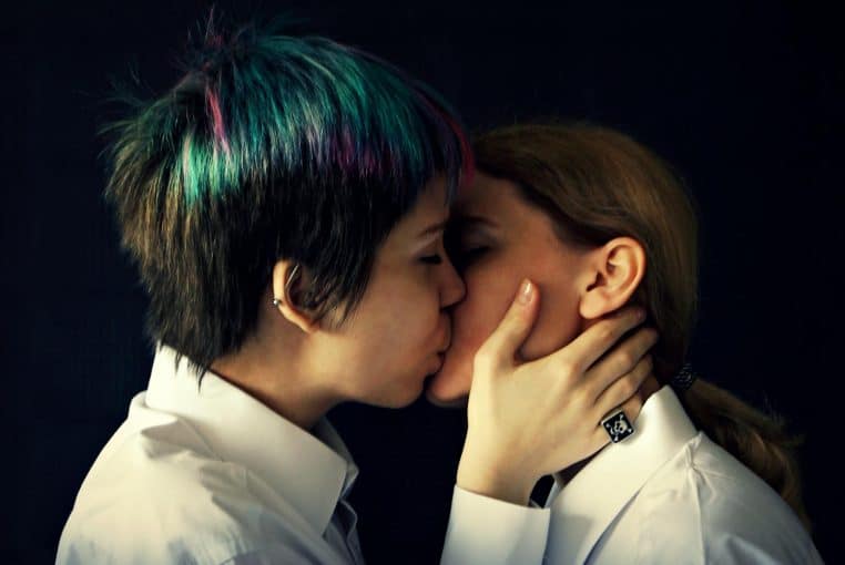 Close-up shot of two women sharing an intimate kiss, dressed in white shirts against a dark background.