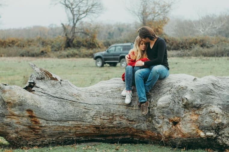 A couple embraces tenderly on a large fallen tree, set against a serene natural landscape.