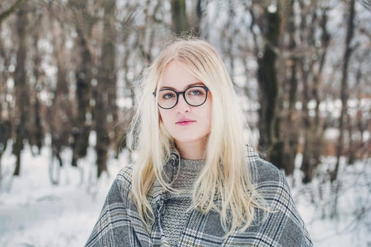 Stylish blonde woman with glasses in a snowy forest, winter fashion.