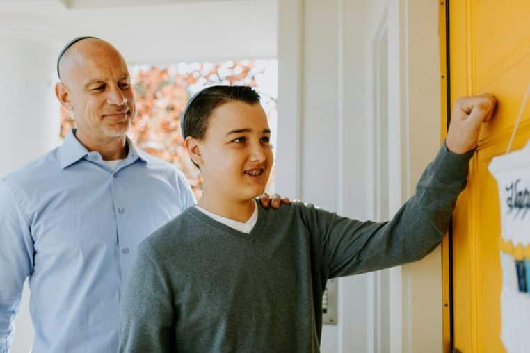 Father and son in traditional Jewish attire knocking on a door for a holiday celebration.